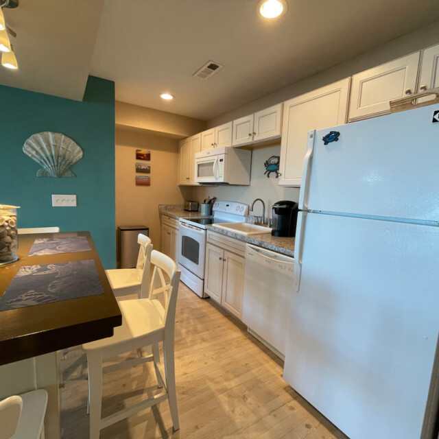 A small kitchen with white cabinets, a white fridge, stove, microwave, and a wooden dining table with chairs.
