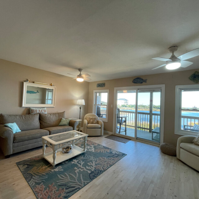 Living room with beige sofas, ceiling fans, and large windows overlooking a waterfront balcony on a sunny day.