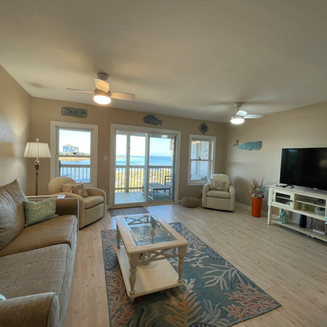 Living room with sofa, chairs, TV, coffee table, and balcony doors overlooking the ocean under a clear sky.