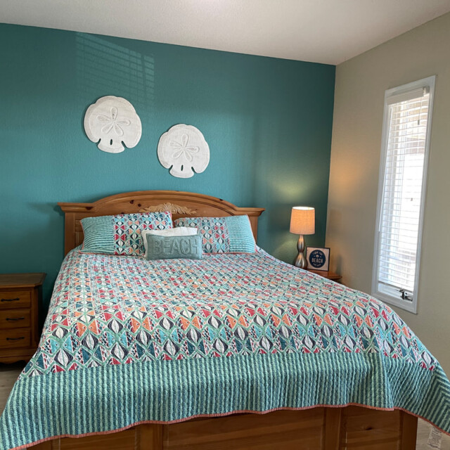 Bedroom with a wooden bed, colorful quilt, bedside tables, lamp, and beach-themed decor on teal and tan walls.