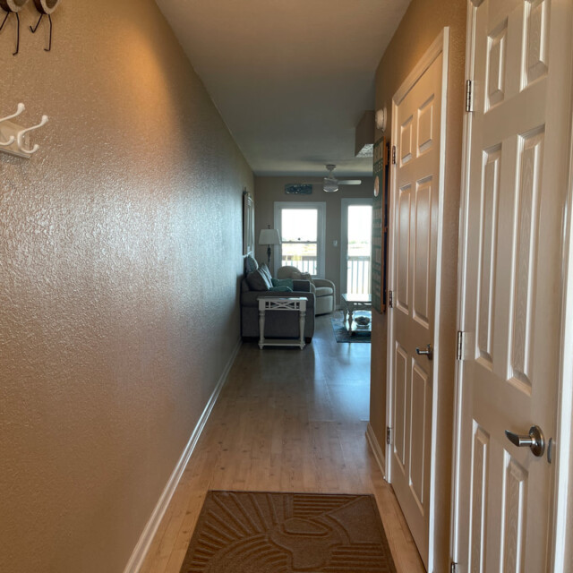 View down a hallway with tan walls, wood floor, rug, and an open door leading to a living room with windows.