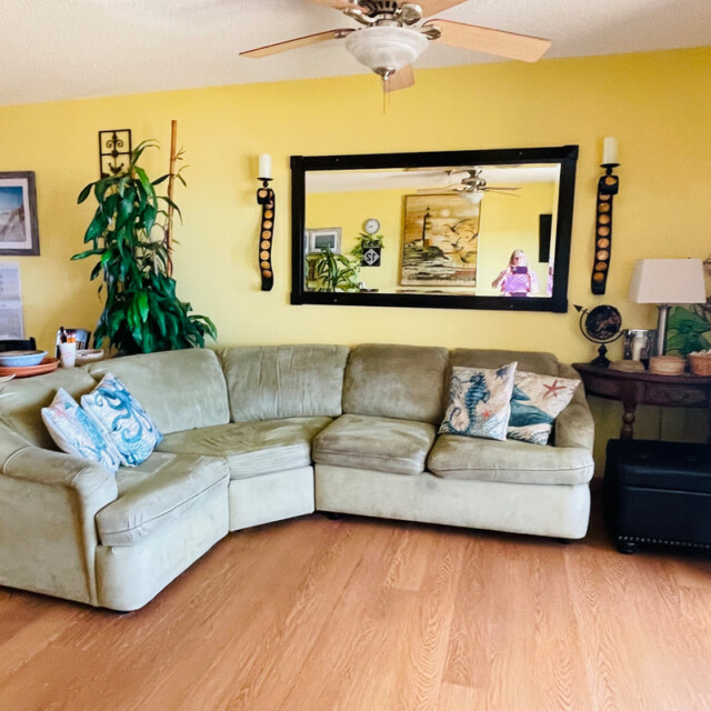 Living room with a green sectional sofa, plants, wall art, mirror, wooden floor, and a ceiling fan.
