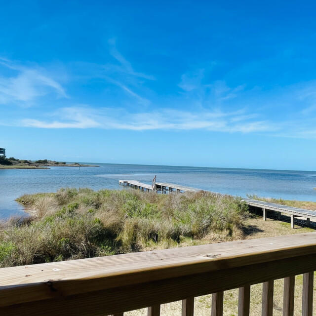 View of a wooden deck overlooking marsh grass, a dock, and calm water under a clear blue sky.