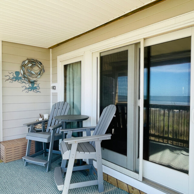 Two gray chairs and a small table on a balcony with sliding glass doors and ocean view in the background.