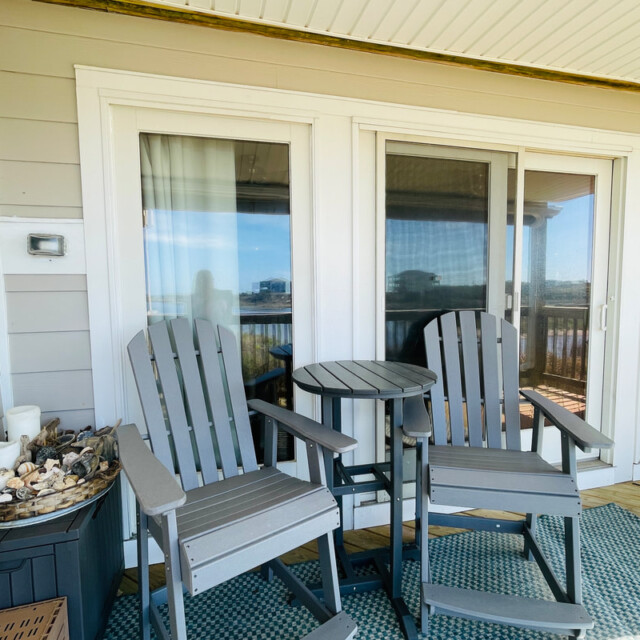 Two gray patio chairs and a small round table on a porch, with seashells and candles on a side table nearby.