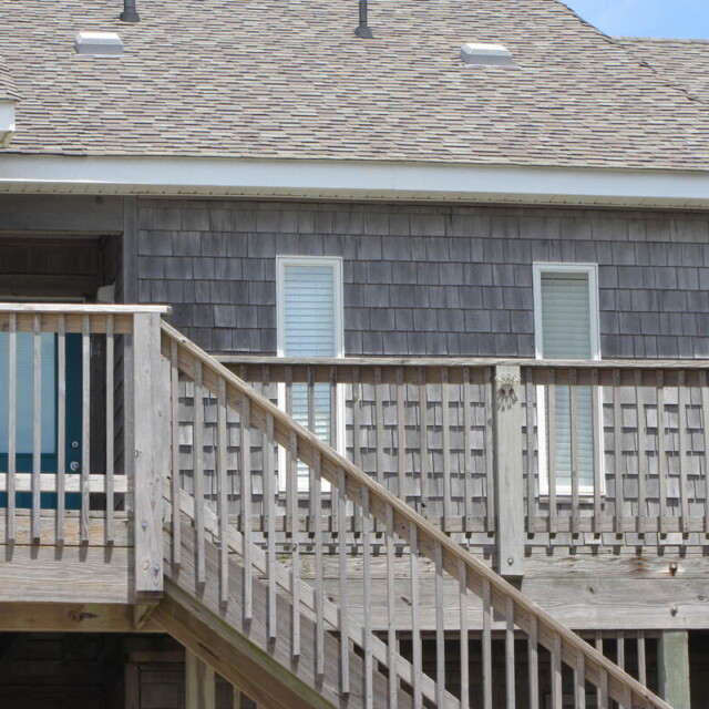 Wooden stairs and railing lead up to a beach house with gray shingles and two windows with closed blinds.