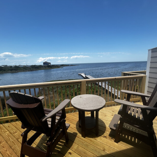 Two wooden chairs and a table on a deck overlook a calm body of water under a clear, sunny sky.