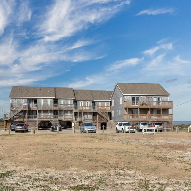 Three two-story beach houses with parked cars in front, under a blue sky with light clouds.
