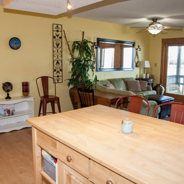 Open living room with a sofa, chairs, plants, and a view of a patio through glass doors, seen from a kitchen island.