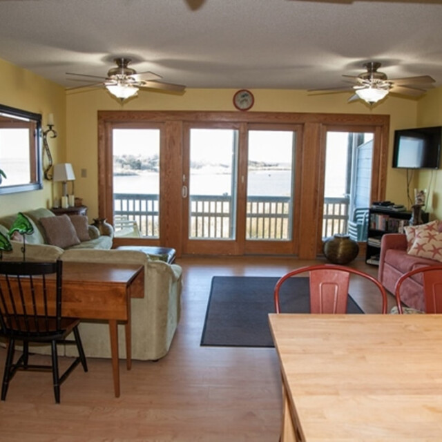 Living room with sofas, chairs, large windows overlooking water, plants, and ceiling fans, decorated in warm tones.
