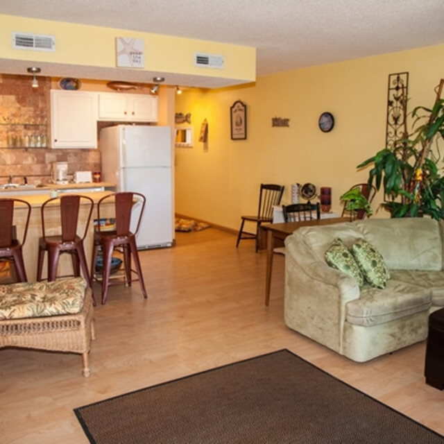 Cozy living room with a green sectional sofa, wicker chair, and a small kitchen with barstools in the background.