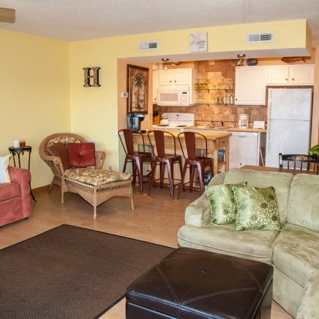 Cozy living room with assorted seating, a brown rug, and a view of a kitchen with barstools and appliances.