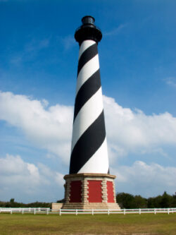 Tall lighthouse with black and white spiral stripes, brick base, blue sky, and white fence in the background.
