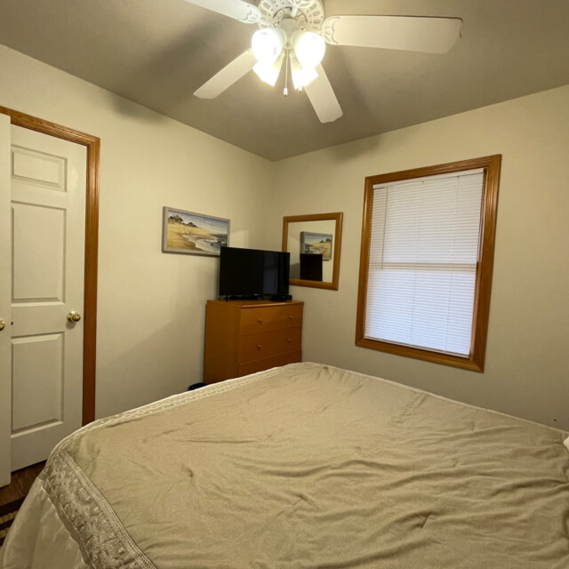 Small bedroom with a bed, dresser, TV, window with blinds, ceiling fan, and framed pictures on cream-colored walls.