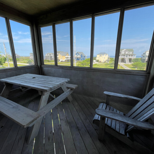 Screened porch with a wooden picnic table and chair, overlooking houses and a blue sky through large windows.