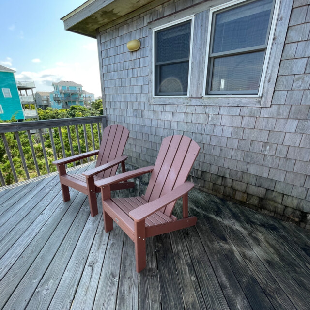 Two wooden chairs sit on a weathered deck outside a shingle-sided house, with other houses visible in the background.
