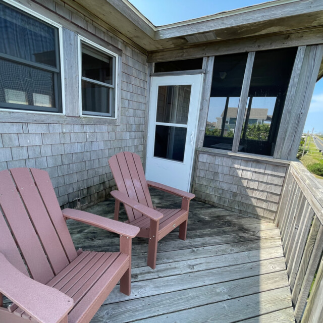 Two wooden chairs sit on a weathered balcony with a view of a residential street and houses in the background.