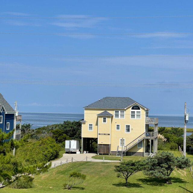 A yellow house on stilts sits near the coast, surrounded by greenery, with blue sky and ocean in the background.