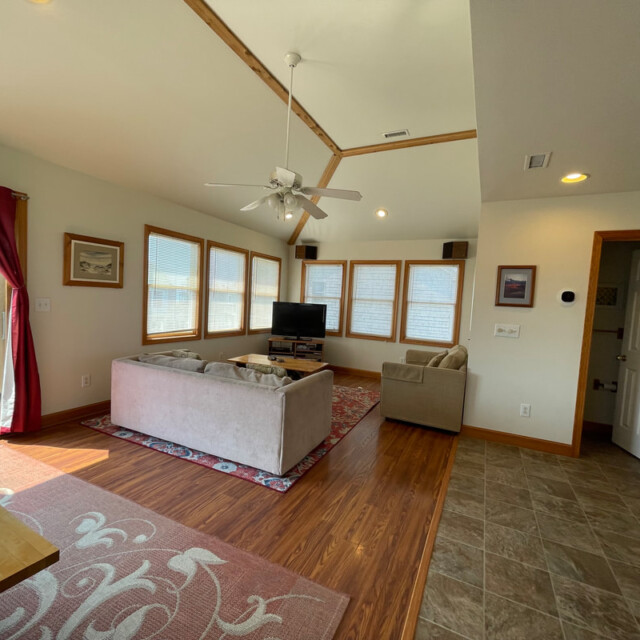 Living room with two beige sofas, TV, ceiling fan, large windows, and wooden floors next to a tiled dining area.