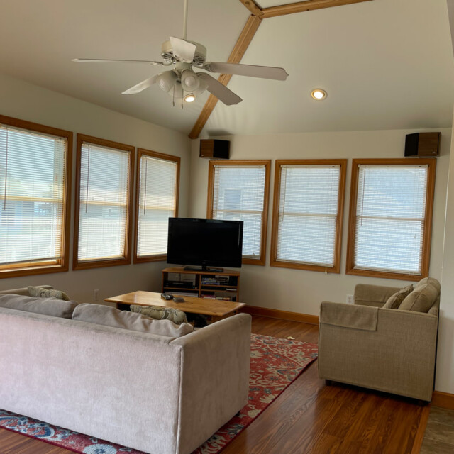 Living room with beige couches, a TV on a stand, wood floors, large windows, ceiling fan, and framed wall art.