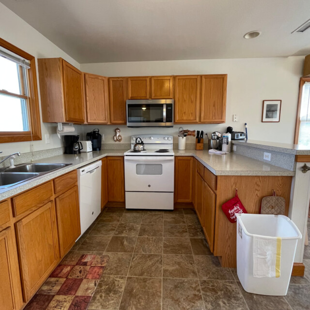 A kitchen with wooden cabinets, white appliances, a trash bin, coffee makers, and a window above the sink.