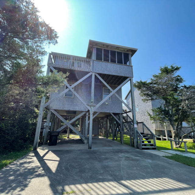 A stilted wooden beach house with a balcony, surrounded by trees on a clear sunny day.