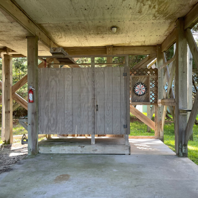 Outdoor wooden shower stall under a raised deck, with a fire extinguisher and dartboard mounted nearby.