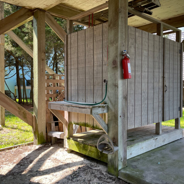 Outdoor wooden shower stall with a hose and fire extinguisher attached to the exterior, under a raised building.