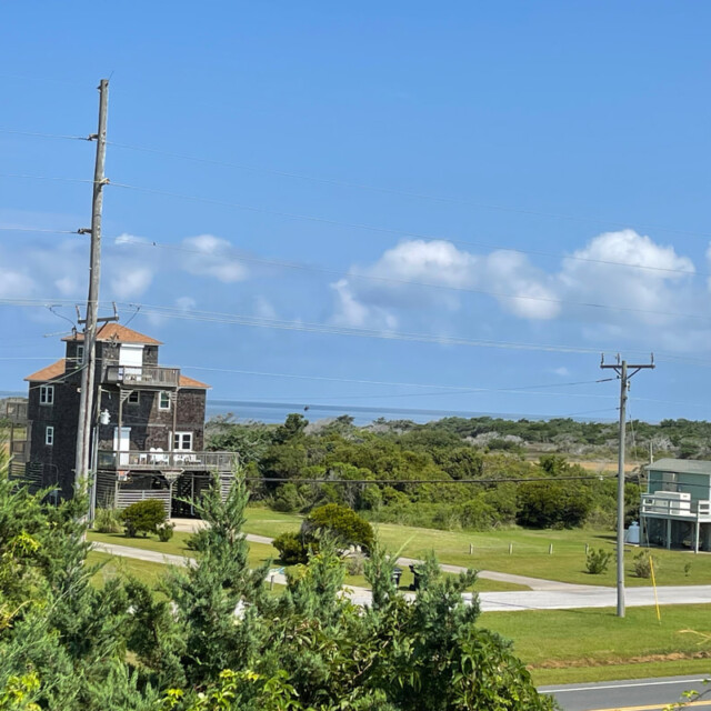 Two houses on grassy land with power lines, trees, and a view of the ocean under a partly cloudy blue sky.