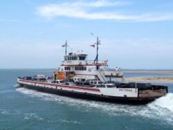 Car ferry named Hatteras crossing water with vehicles on board, against a backdrop of sky and sandy shore.