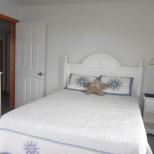 A white bed with star-themed pillows, a nightstand with a lamp, and a dresser with flowers in a bright bedroom.
