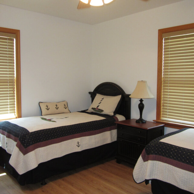 Bedroom with two twin beds, wood blinds, nautical-themed bedding, nightstand, and lamp on wooden floor.