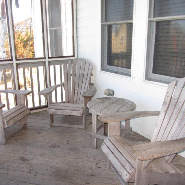 Three wooden Adirondack chairs and a small round table on a screened-in porch with wooden floor and white walls.