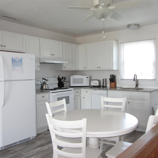 White kitchen with refrigerator, stove, microwave, and round dining table with four chairs. Window above sink.