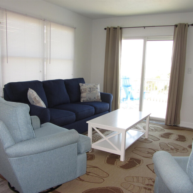 Living room with blue sofa, two blue chairs, white coffee table, and large windows with beige curtains and blinds.