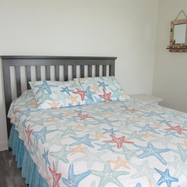 Bed with a starfish-patterned quilt and pillow, wooden headboard, white walls, small mirror and shelf on the wall.