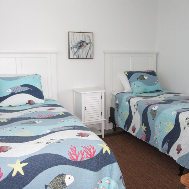 Two twin beds with fish-patterned bedding and a small white nightstand between them in a simple, white room.
