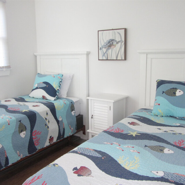 Two twin beds with ocean-themed bedding in a white room, separated by a small white nightstand and wall art above.