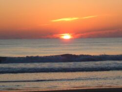 Orange sun rises over calm ocean waves with a clear sky and minimal clouds near the horizon.
