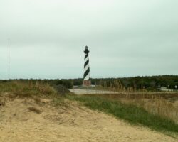 A black and white striped lighthouse stands near a sandy area with grass and a cloudy sky overhead.