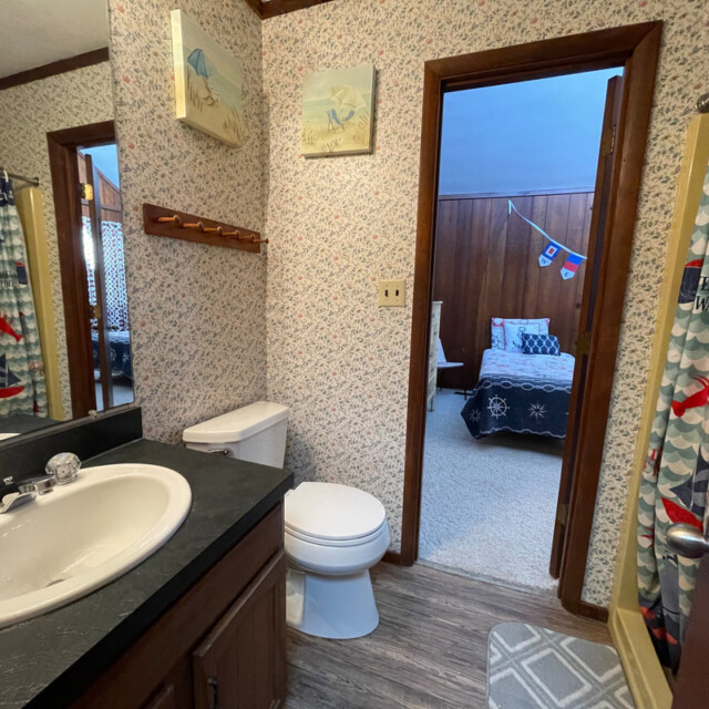 Bathroom with a sink, toilet, nautical shower curtain, and view into a bedroom with wood-paneled walls and bed.