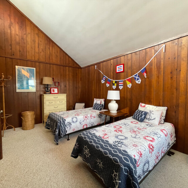 Wood-paneled bedroom with two twin beds, nautical decor, dresser, lamp, and coat rack on beige carpeted floor.