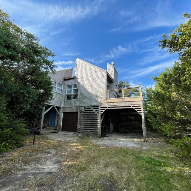 Two-story wooden house with a deck, surrounded by trees, under a blue sky with scattered clouds.