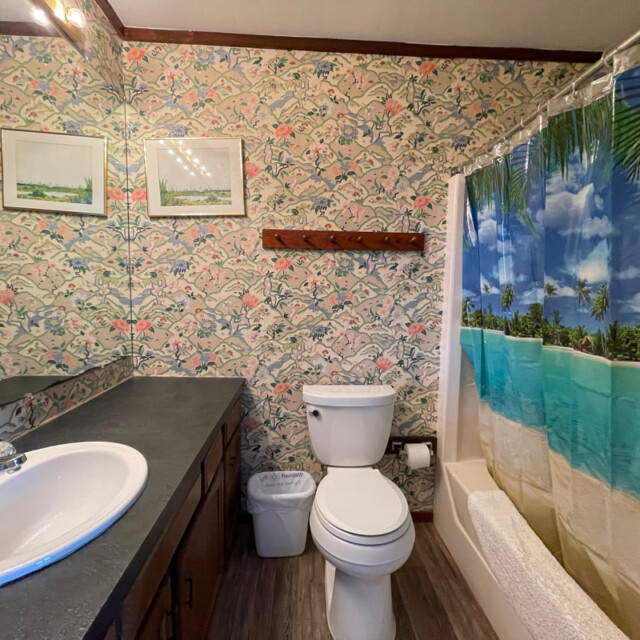 Bathroom with floral wallpaper, a sink, toilet, trash bin, and a bathtub with a beach-themed shower curtain.