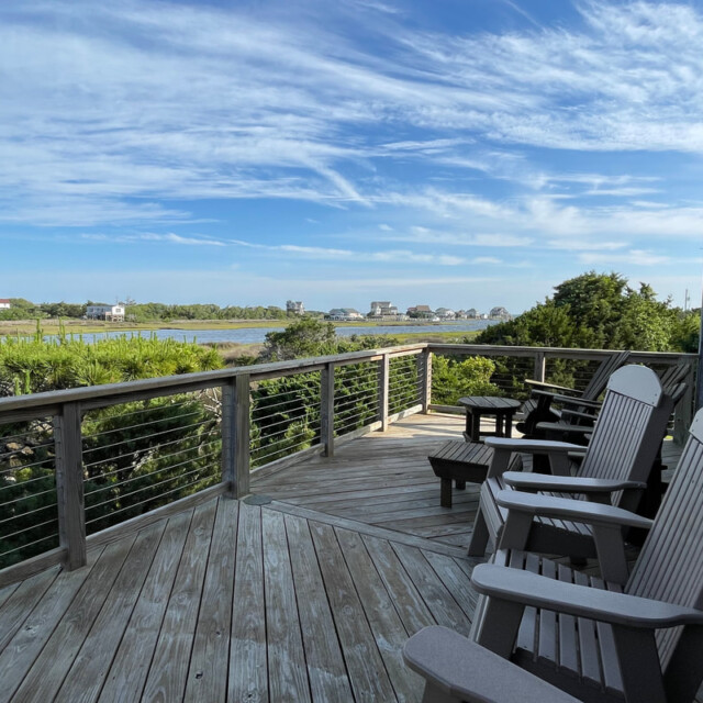 Wooden deck with Adirondack chairs overlooks greenery, water, and distant houses under a blue sky with wispy clouds.