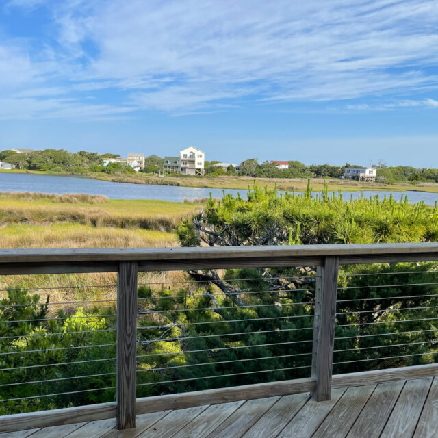 Wooden deck with railing overlooks grassy marsh, water, and distant houses under a blue sky with scattered clouds.