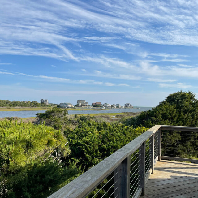 Wooden deck overlooks green shrubs, water, and distant houses under a blue sky with scattered clouds.