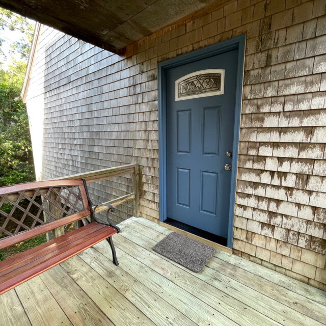 Wooden porch with a blue door, brown bench, welcome mat, and shingle siding next to trees.