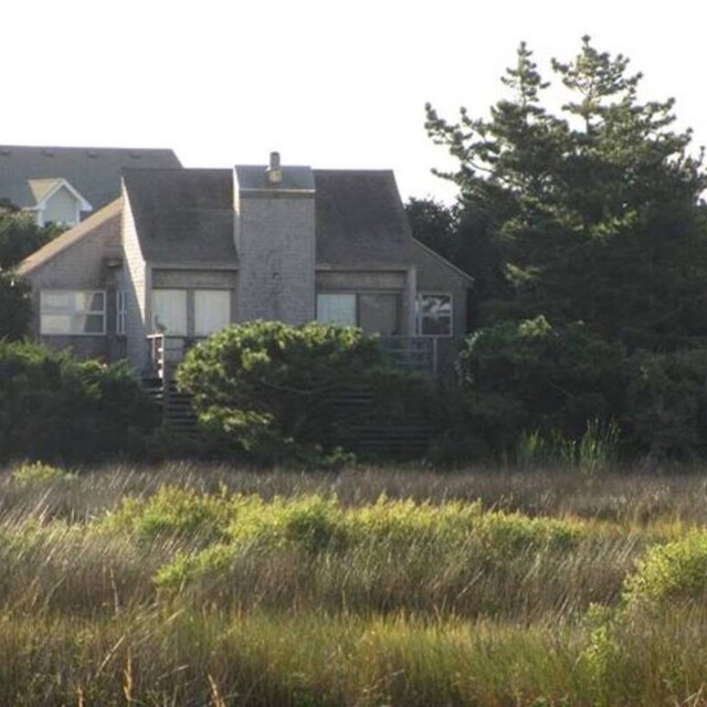 A small, gray, weathered house sits among trees and tall grass with other houses nearby in the background.
