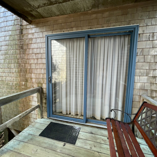 A wooden bench and doormat on a small deck in front of a sliding glass door with closed curtains.